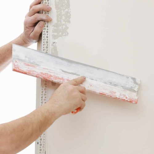 Builder in protective mask using a trowel to add plaster. Plastering wall with putty-knife, close up